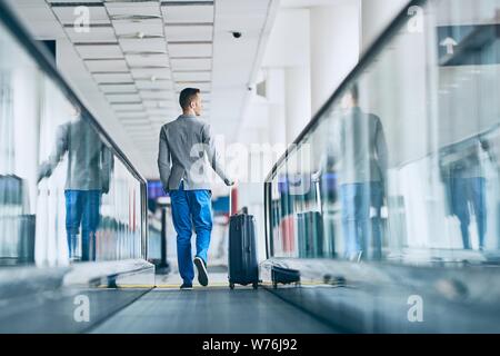 Well dressed young man travel by airplane. Businessman with luggage walking on travelator at airport. Stock Photo