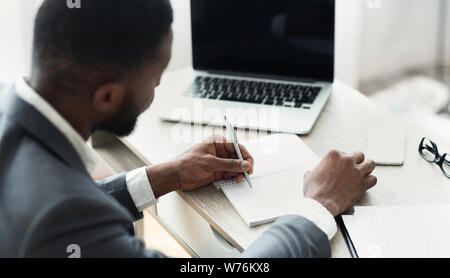 Black businessman working on laptop and taking notes to notepad Stock Photo
