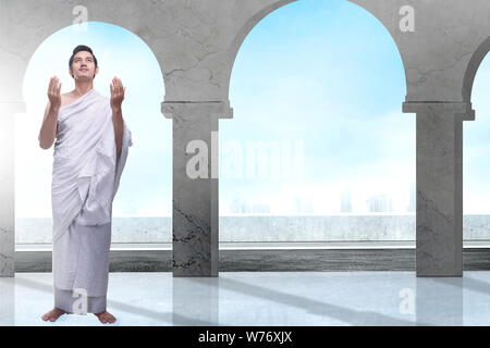 Asian Muslim man in ihram clothes standing and praying while raised arms inside the mosque Stock Photo