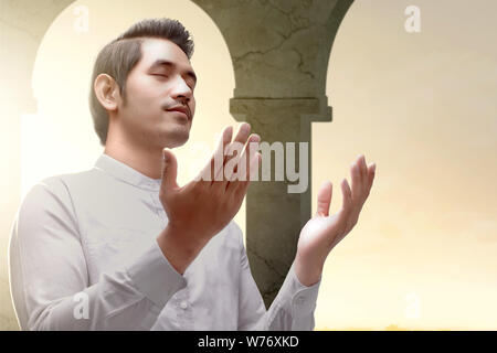 Asian Muslim man standing and praying while raised arms inside the mosque Stock Photo