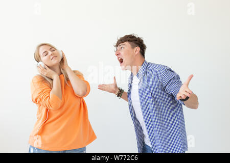 Young upset man begs his woman to listen to him but she listens to music with headphones posing on a white background. Misunderstanding and Stock Photo