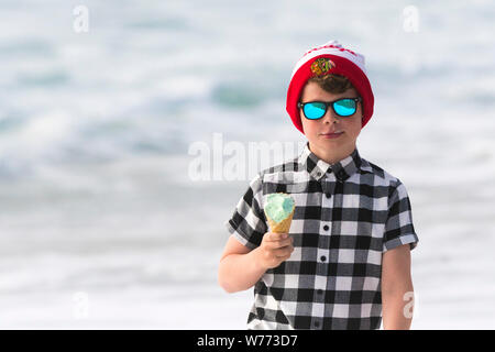 A young boy wearing reflective sunglasses, check shirt and a Chicago Blackhawks bobble hat eating an ice cream. Stock Photo