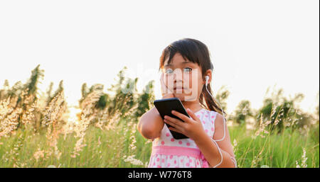 Asian child girl Listening to music and singing from a mobile phone and happy on meadow in summer in nature Stock Photo