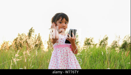 Asian child girl Listening to music and singing from a mobile phone and happy on meadow in summer in nature Stock Photo