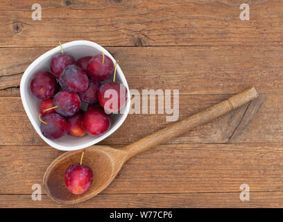 Red cherry plums in bowl, with wooden spoon, on wooden background. Stock Photo