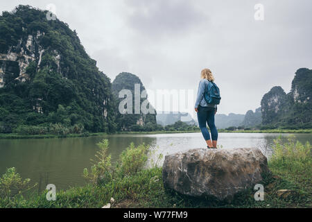 Caucasian blonde woman overlooking limestone mountains in Ninh Binh province, Vietnam. Cloudy day, view from the back, reflection in water Stock Photo