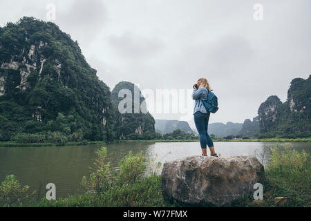 Caucasian blonde woman taking pictures of limestone mountains in Ninh Binh province, Vietnam. Cloudy day, view from the back, reflection in water Stock Photo