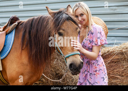 Young blonde woman smiling and stroking a brown horse before a walk that eats hay near the haystack on a summer clear day Stock Photo