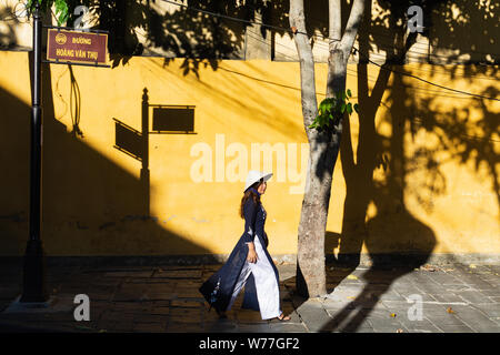 Hoi An, Vietnam - June 2019: Vietnamese woman walking and dropping shadow on yellow wall in old town district Stock Photo
