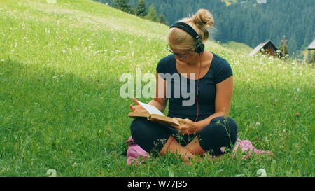 blonde girl reads book on a green grass listening to music with headphones Stock Photo
