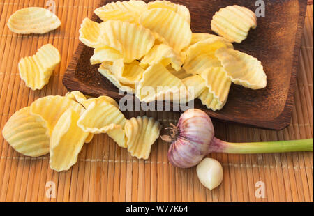 Appetizing crispy potato chips on a woven bamboo napkin, in a wooden bowl, next to the head of garlic, unhealthy or wrong food, but very tasty. Stock Photo