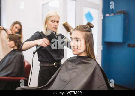 Portrait of happy woman at the hair salon. Professional hair styling concept. Hairdresser drying girl long hair using hairdryer and brush. Drying With Stock Photo