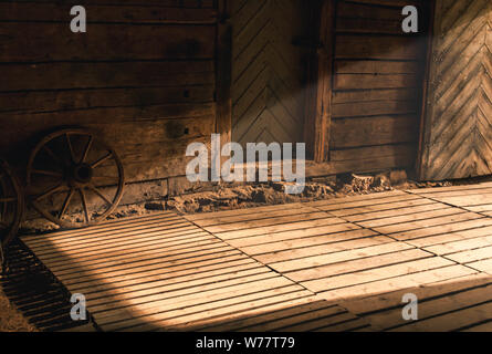 Wooden wheel leaned against a wooden wall with light streaming inside the room from opened doors. Stock Photo