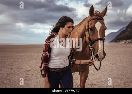 Beautiful woman walking with a brown horse on the coast. Cowgirl taking a walk with her stallion in evening. Stock Photo