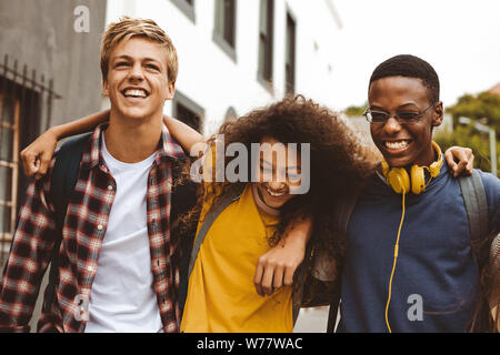Close up of three college friends standing in the street with arms around each other. Cheerful boys and a girl wearing college bags having fun walking Stock Photo