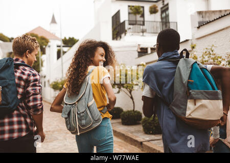 Rear view of teenage friends walking together in the street. Smiling teenage girl walking with friends wearing college bag. Stock Photo