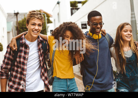 Teenage boys and girls walking in the street holding each other. Smiling college friends walking together in street wearing college bags having fun. Stock Photo