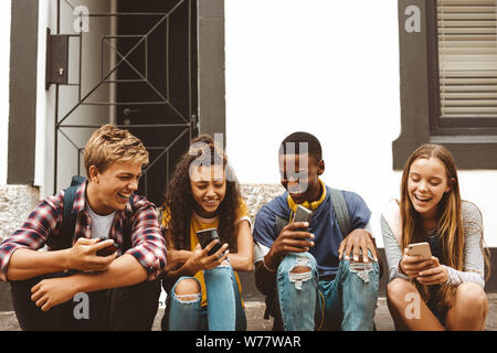 Teenage friends sitting on a pavement holding their mobile phones. Cheerful college boys and girls having fun talking sitting outdoors in a street. Stock Photo