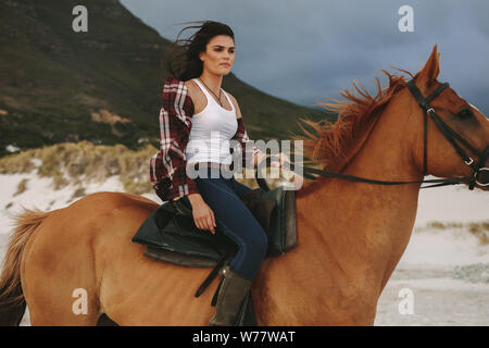 Attractive young woman riding a horse at the beach in evening. Cowgirl riding with her stallion on the sea shore. Stock Photo