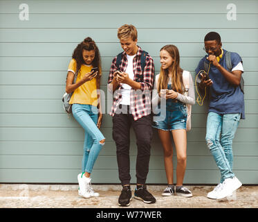 Group of multi ethnic friends standing in the street and looking at their mobile phones. Happy teenage boys and girls wearing college bags talking whi Stock Photo
