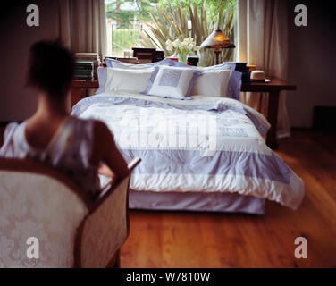 Bed in a room with a woman sitting in front of it on a chair. Stock Photo