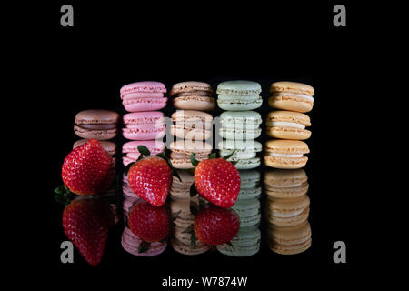Many macaroons stacked next to two juicy red strawberries on a glass table and a black background - Tasty macaroon dessert in different colours - Clos Stock Photo