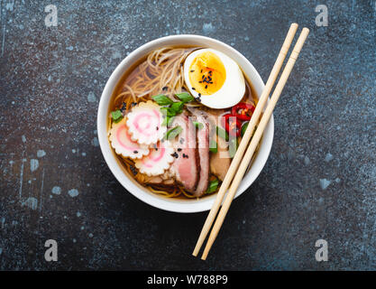 Tasty Japanese noodle soup ramen in white ceramic bowl with meat broth, sliced pork, narutomaki, egg with yolk on rustic stone background. Traditional Stock Photo