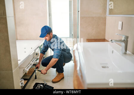 Young worker in uniform sitting on squats while fixing or fastening detail Stock Photo