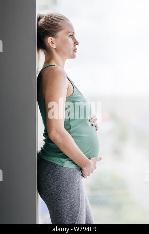 Pregnant woman stands near the window wearing sportswear. Stock Photo