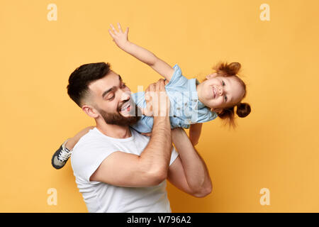 cheerful awesome father lifting his kid, playing with her. little girl pretends to be a plane, girl learning to fly . isolated yellow background. stud Stock Photo