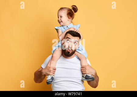 surprised puzzled guy looking up, at his little sister who is sitting on his shoulders . close up photo. isolated yellow background. Stock Photo