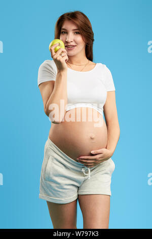 positive woman with long brown hair holding an apple and touching her big belly. close up photo. isolated blue background. studio shot Stock Photo