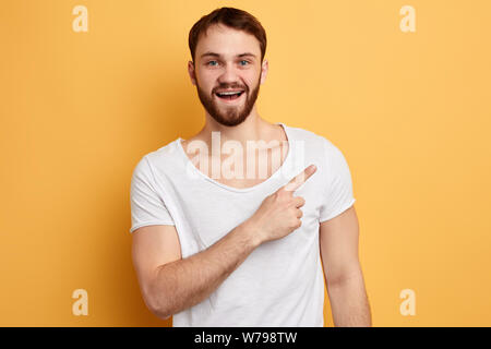 happy handsome young man in white t-shirt pointing finger away at copy space isolated over yellow background. close up photo. direction concept. this Stock Photo