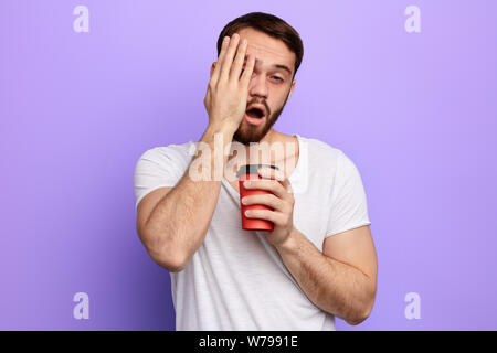 sleepy handsome man with a palm on his cheek drinking coffee in the morning, difficult monday, man cannot wake up, close up portrait isolated blue bac Stock Photo