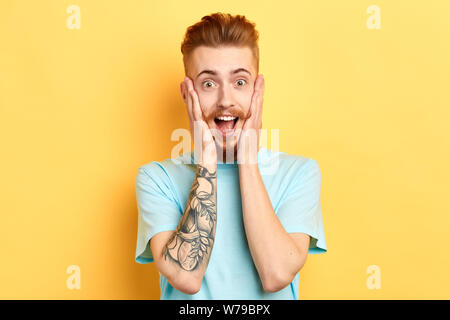 stunned terrified young attractive man with funny moustache, beard, covers mouth and looks with eyes popped out down, being shocked by gossip, news. o Stock Photo
