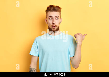 emotional funny man looking aside, being horrified to see something, indicates with thumb aside, isolated over yellow background. close up portrait. c Stock Photo