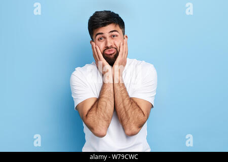 stressed,tired, crying handsome man squeezing cheeks with palms, being indifferent, annoyed and bored, standing over blue background. close up portrai Stock Photo