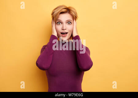 Beautiful young fair-haired woman with palms on her head and wide open mouth over isolated yellow background.. isolated yellow background. studio shot Stock Photo