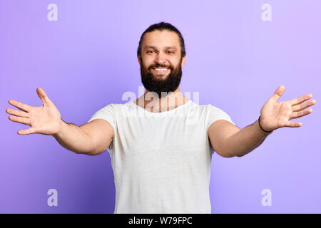 Young handsome cheerful man wearing white t-shirt greeting guests, hugging them over isolated blue background .Cheerful expression, happiness.feeling Stock Photo