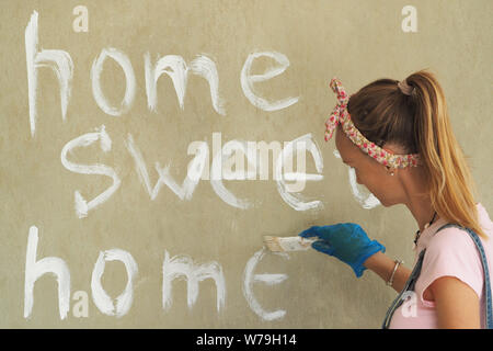 Beautiful woman with a brush and roller for painting. Background gray cement wall with the inscription home sweet home. Stock Photo