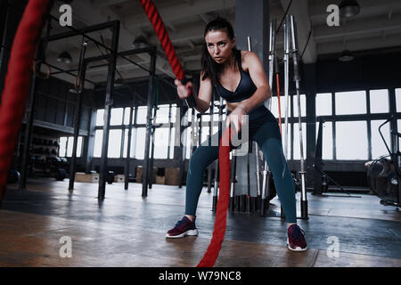 Fit woman using battle ropes during strength training at the gym. Athlete moving the ropes for burning fats at health club. Stock Photo