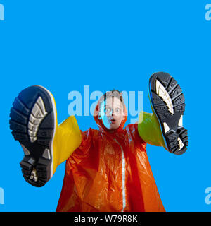 smiling boy in orange raincoat stuck his hands in rubber boots on a blue background Stock Photo