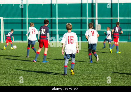 Boys in red and blue sportswear plays  football on field, dribbles ball. Young soccer players with ball on green grass. Training, football Stock Photo