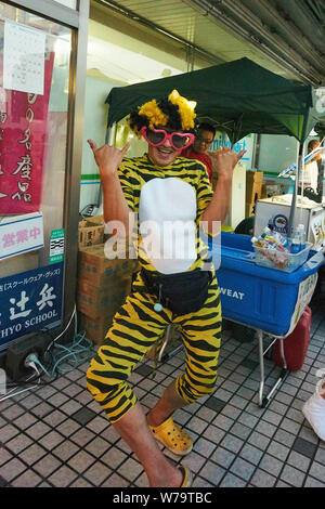 Man dressed in bright yellow tiger-print body suit, heart sunglasses, and a fanny pack poses with shakas at the Akita Kanto Matsuri festival in 2016. Stock Photo