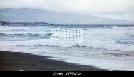 Powerful ocean waves crashing on wet beach under cloudy sky with island in background, in Azores, Portugal.