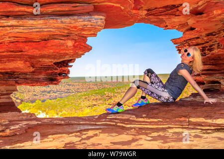 Happy woman sitting inside the red rock arch in rugged sandstone of Nature's Window, Kalbarri National Park. Caucasian blonde girl enjoying in Western Stock Photo