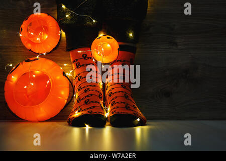 kids legs in stylish warm bright colorful striped funny socks in garland lights on floor with pumpkins in room. decor for Halloween, cosy moment. Stock Photo