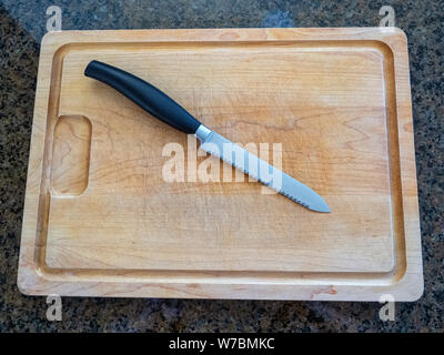 Top view of an Isolated serrated utility knife on a large wooden cutting board with a granite kitchen counter top background Stock Photo