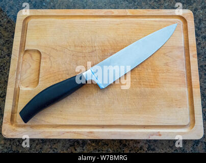 Isolated large chef’s knife on a large wooden cutting board with a granite kitchen counter top background Stock Photo