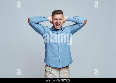 Bla bla bla. Depressed man closing ears with both hands and screaming while standing against grey background. Stress emotion. Noise concept Stock Photo